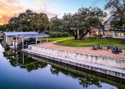 Private waterfront backyard with boat dock, native trees, and a firepit lounge area overlooking the lake at sunset.