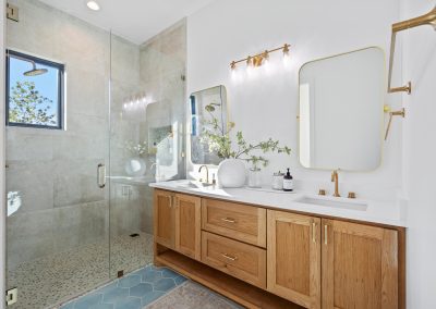 Double vanity bathroom with oak cabinetry, walk-in shower, pebbled tile flooring, and brushed brass plumbing and lighting fixtures.