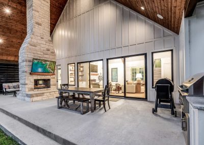 Covered outdoor patio with built-in grill, stone fireplace, and dining table—framed by vaulted wood ceiling and expansive rear windows.