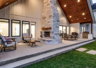Twilight patio with vaulted wood ceiling, outdoor stone fireplace, grill station, dining area, and cushioned lounge seating.