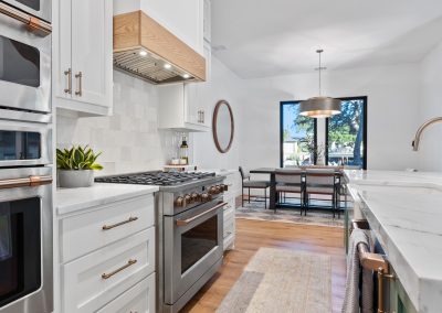 Kitchen with white cabinetry, marble countertops, wood range hood, double ovens, and open flow to dining area.