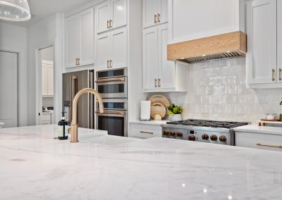 Close-up of kitchen with marble countertops, wood range hood, brushed brass faucet, and glazed tile backsplash.
