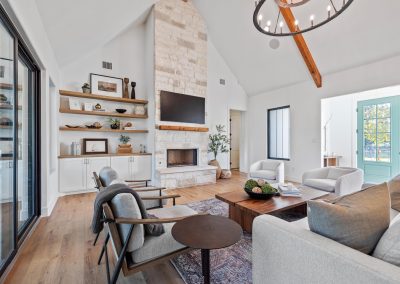 Living room with vaulted ceiling, stone fireplace, wood beams, built-ins, and natural light from large glass doors and windows.