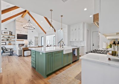 Open-concept kitchen with sage green island, vaulted living room, stone fireplace, wood beams, and white cabinetry.