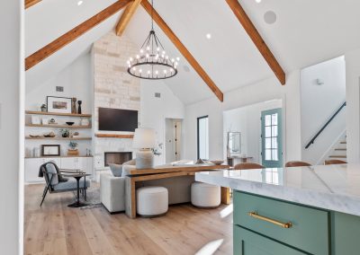 Vaulted ceiling with exposed wood beams above an open kitchen and living area featuring a sage green island and stone fireplace.