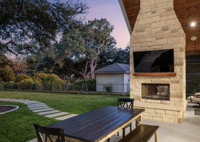 Outdoor dining area with wood-beamed ceiling, stone fireplace, TV, and twilight ambiance overlooking a lush backyard.