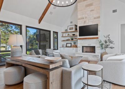 Living room with vaulted ceiling, exposed wood beams, circular chandelier, large windows, and a full-height stone fireplace.
